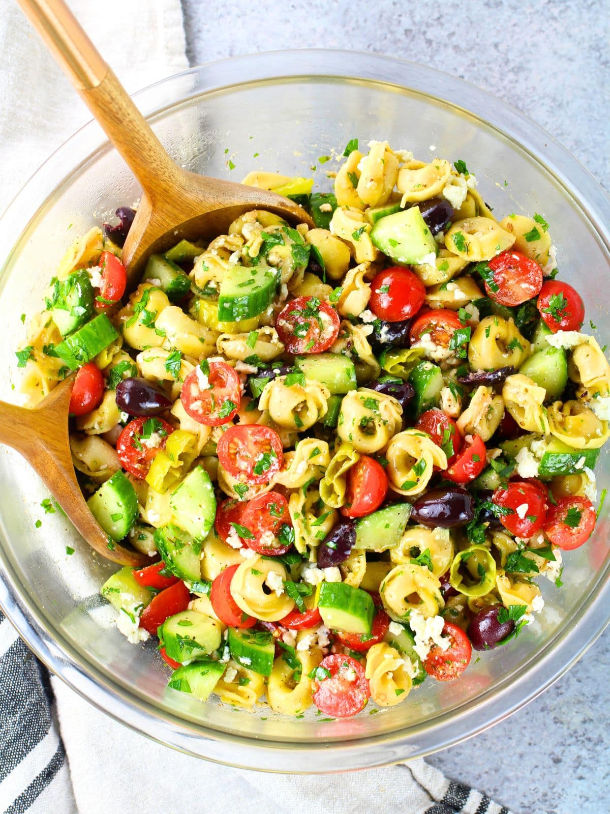 Greek tortellini pasta salad with cherry tomatoes, cucumber, Kalamata olives, and feta in a glass bowl with a wooden spoon