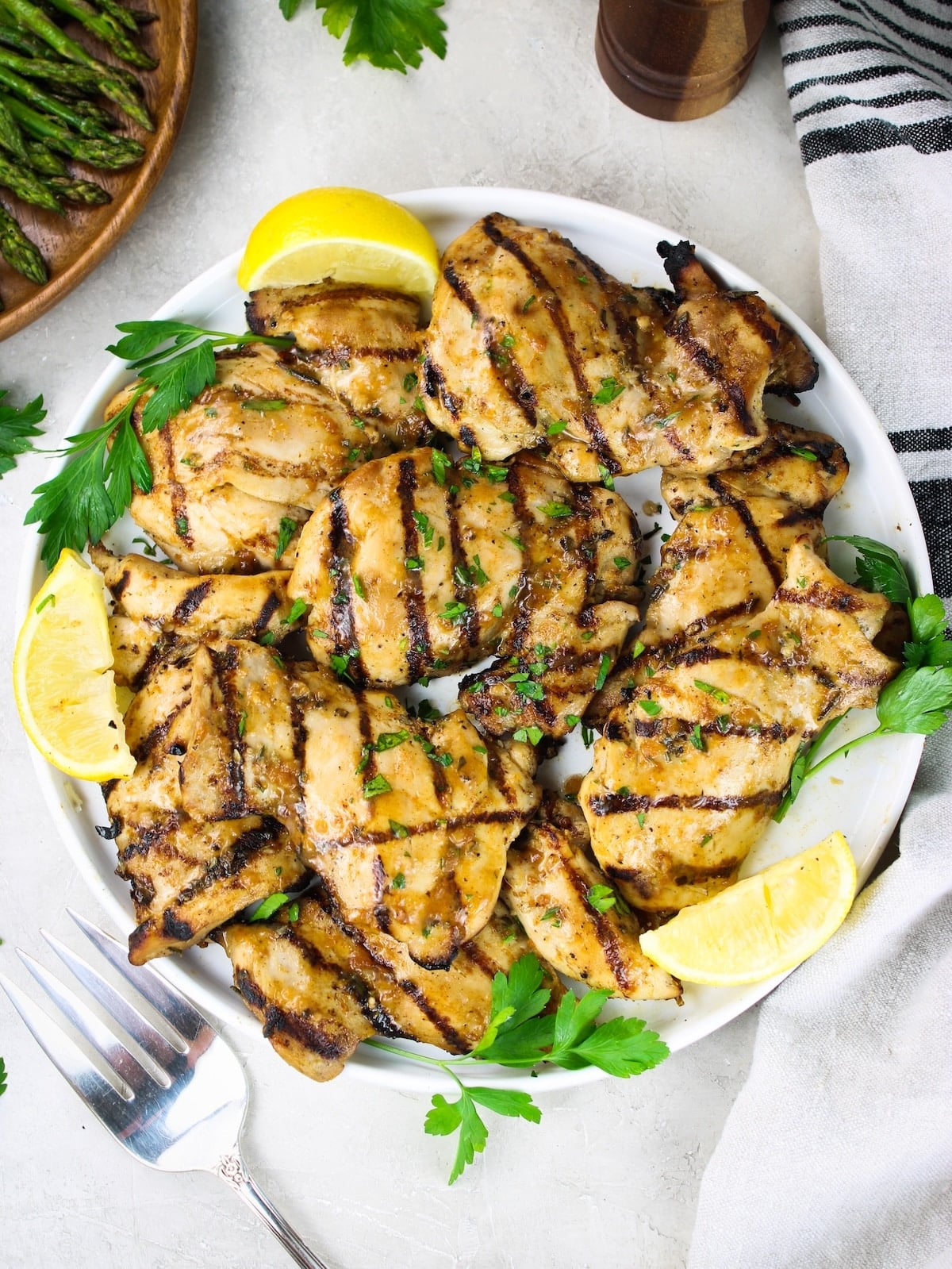 Overhead view of a white plate of grilled chicken thighs garnished with lemon wedges, fresh parsley, and asparagus on the side