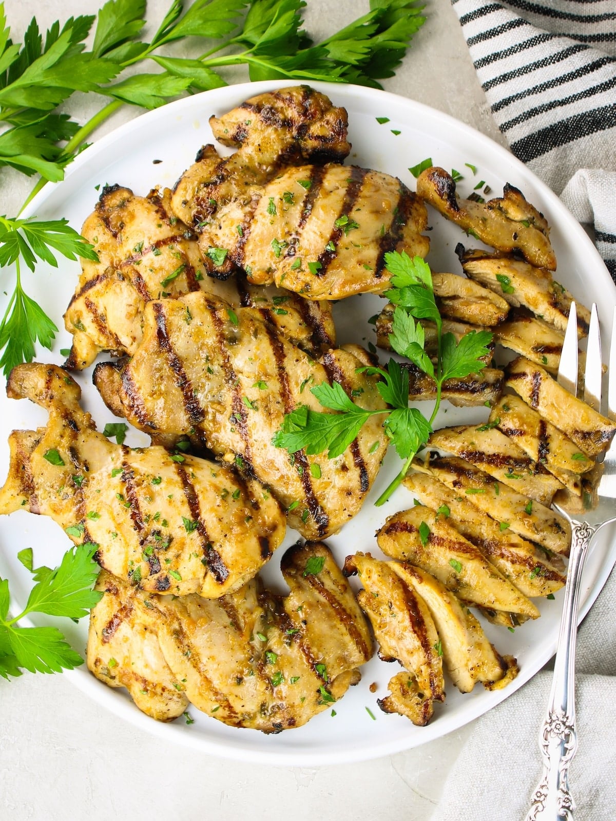 Overhead view of grilled boneless chicken thighs on a white plate, one sliced to show juicy interior, garnished with fresh parsley