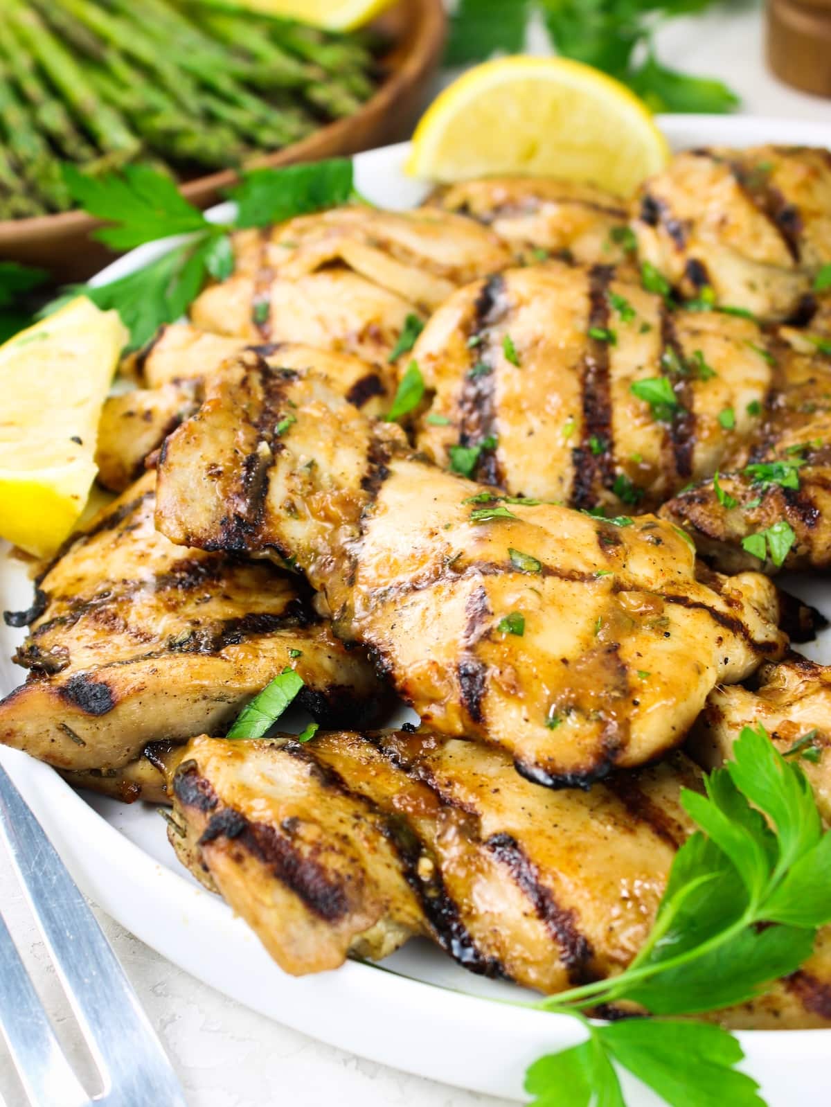 Platter of caramelized grilled chicken thighs with lemon wedges, fresh parsley, and asparagus in the background