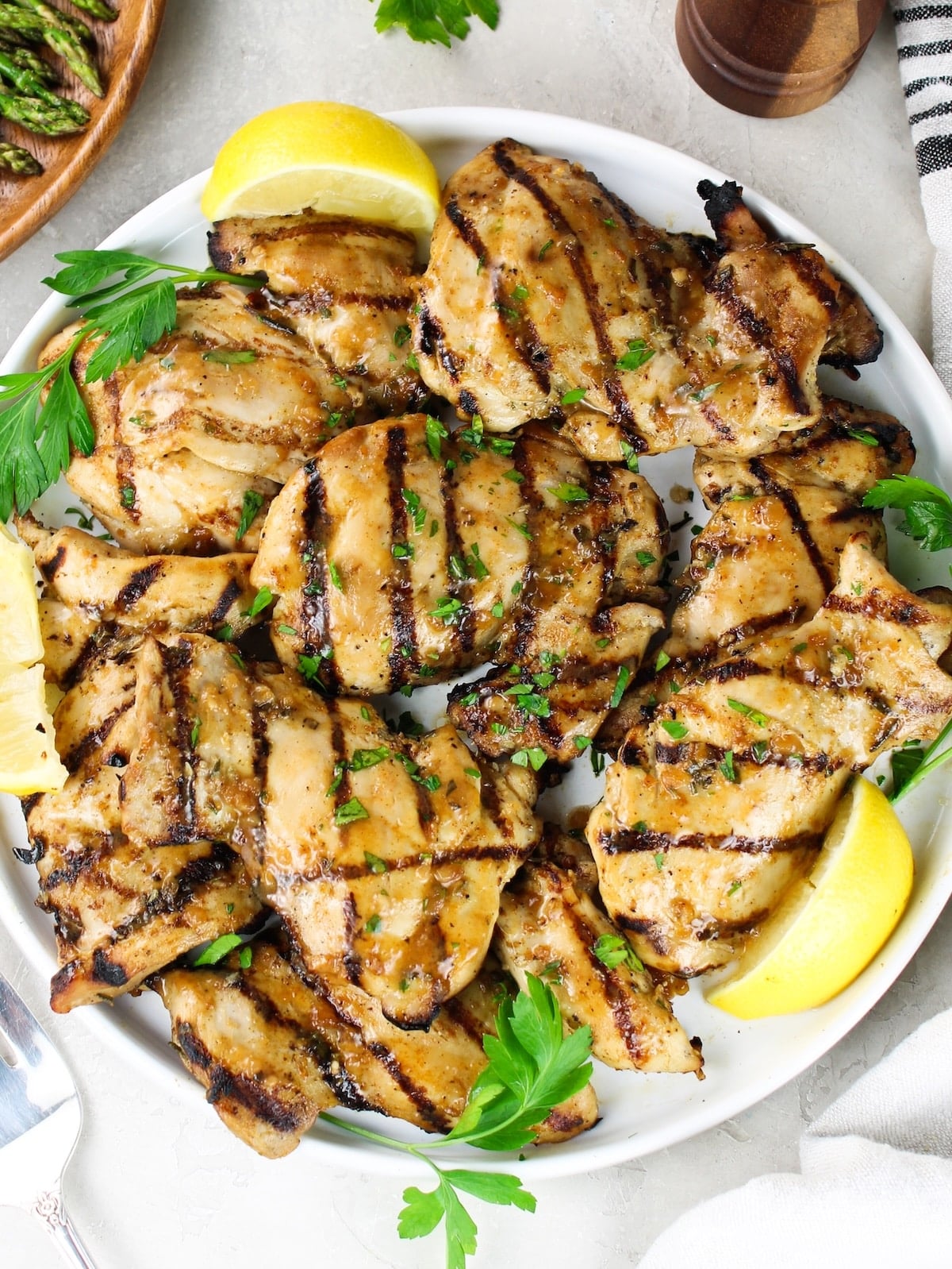 Grilled chicken thighs with caramelized grill marks on a white serving plate with lemon wedges, parsley, and a fork