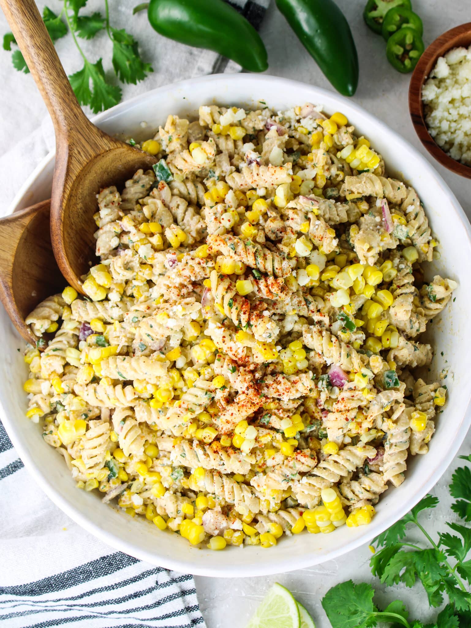 Overhead shot of Mexican street corn pasta salad with two wooden spoons and a bowl of cotija cheese