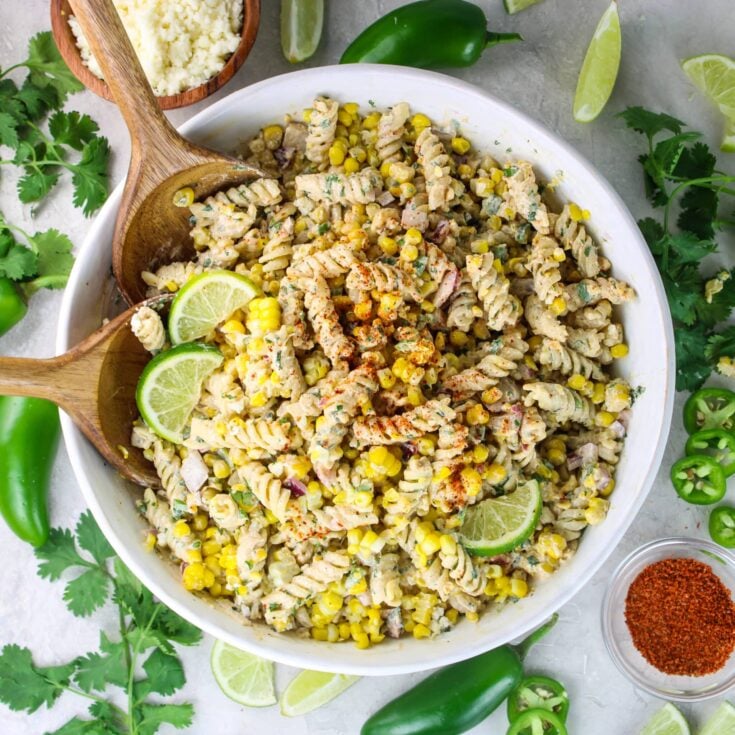 Square shot of elote pasta salad in a white bowl surrounded by lime wedges and jalapeño