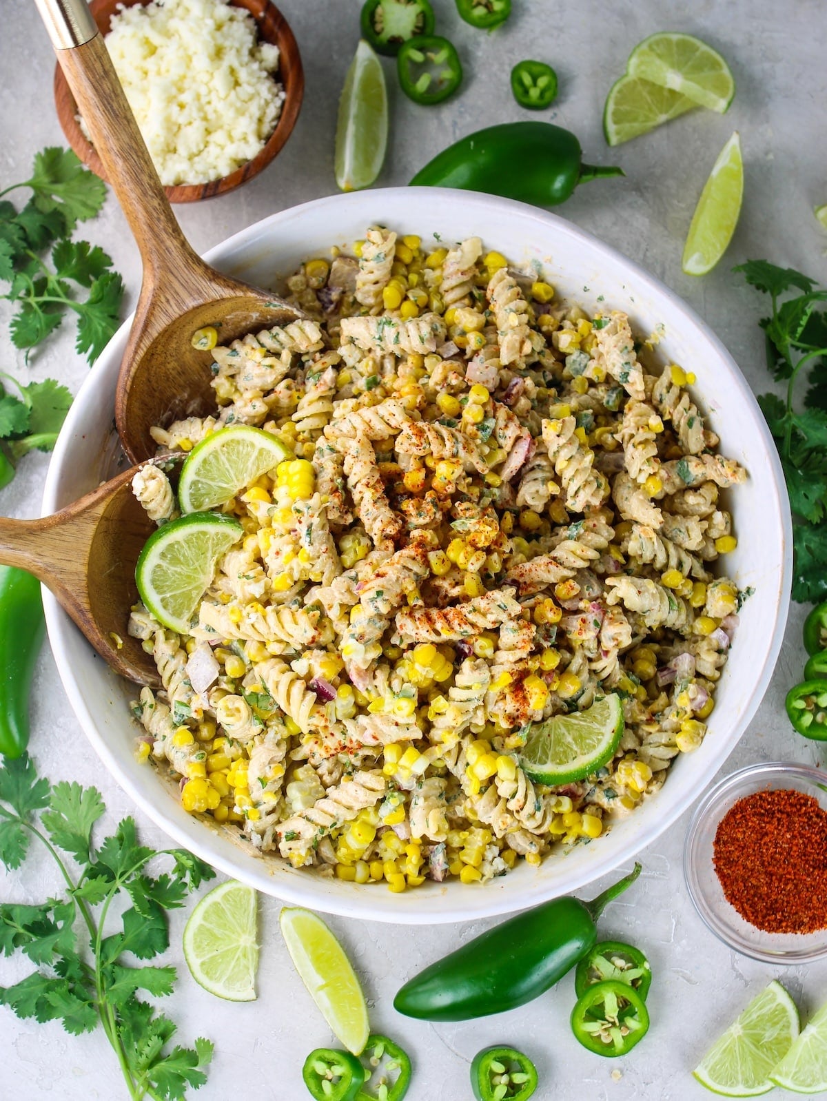 Mexican street corn pasta salad in a white bowl surrounded by lime wedges, jalapeño, cotija cheese, and tajin