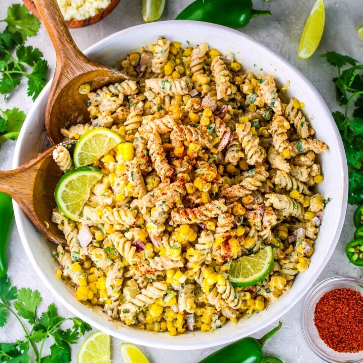 Mexican street corn pasta salad in a white bowl with fresh lime wedges, jalapeño, and Tajin and cotija cheese on the side.